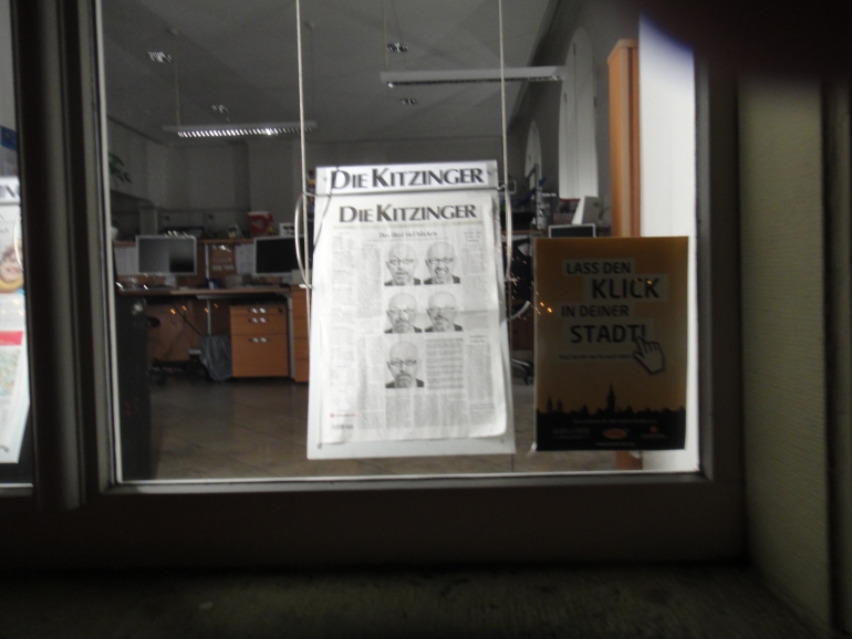 Screenshot vom Schaufenster der Mainpost-Filiale. Der Banner der Kampagne "Lass den Klick in deiner Stadt" ist am Glas angebracht, dahinter wird die aktuelle Ausgabe der Kitzinger Zeitung ausgestellt.